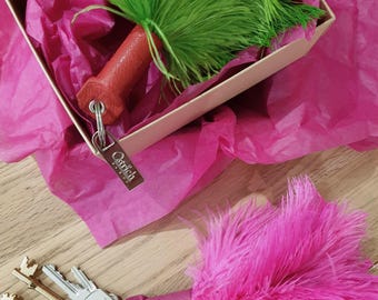 Puede incluir: Dos llaveros con plumas de avestruz en verde vibrante y rosa brillante unidas a mangos de cuero texturizado. Cada llavero incluye una etiqueta de metal plateado grabada con la palabra Ostrich y una argolla con varias llaves plateadas y doradas.