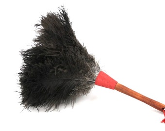 Puede incluir: Un plumero de plumas de avestruz negras con un mango de madera rojo. El mango tiene un cordón rojo en el extremo.