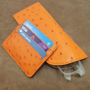 May include: An orange textured card holder and a matching orange glasses case. The card holder contains two cards, one labeled Visa and John Lewis, and the other labeled Revolut. The glasses case holds a pair of clear-framed glasses inside.