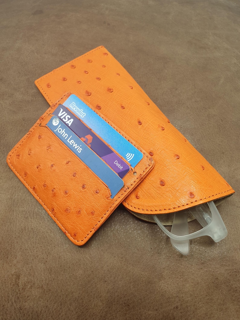 May include: An orange textured card holder and a matching orange glasses case. The card holder contains two cards, one labeled Visa and John Lewis, and the other labeled Revolut. The glasses case holds a pair of clear-framed glasses inside.