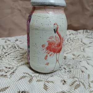 May include: A white glass jar with a pink flamingo painted on the front. The jar has a white lid and is decorated with white speckles.