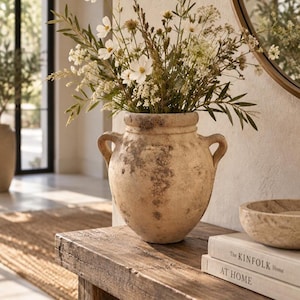 Può includere: Un vaso in terracotta beige usurato con due manici laterali, caratterizzato da una superficie ruvida e maculata. Il vaso è riempito con un bouquet di fiori cosmos bianchi e fogliame verde. Accanto ci sono due libri intitolati The Kinfolk Home.