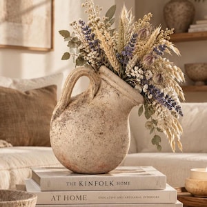 Puede incluir: Una jarra de cerámica beige de textura rústica con una sola asa, llena de un arreglo floral seco de lavanda, trigo y eucalipto. La jarra descansa sobre una pila de tres libros titulados The Kinfolk Home, At Home y The Kinfolk Garden.
