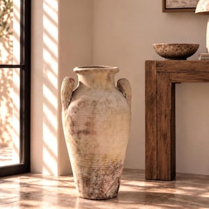 Pode incluir: Um vaso ânfora grande e alto de terracota desgastada com uma borda larga e duas alças laterais. A superfície apresenta um acabamento terroso e manchado em tons de bege, creme e castanho, com sinais de envelhecimento e textura rústica.