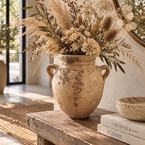 Pode incluir: Um vaso ânfora de cerâmica bege desgastada com duas alças laterais, preenchido com um grande arranjo de gramíneas secas, flores silvestres e vagens. Ao lado do vaso estão dois livros empilhados intitulados The Kinfolk Home e At Home.
