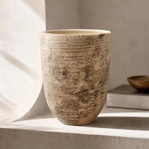 Puede incluir: Un jarrón cilíndrico de cerámica con base cónica, que presenta una superficie texturizada con estrías horizontales y un acabado desgastado y moteado en tonos beige, crema y marrón oscuro, mostrando un aspecto envejecido en el exterior.