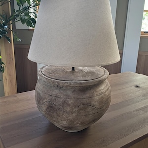 Könnte beinhalten: Ein beigefarbener Stofflampenschirm sitzt auf einem strukturierten, runden Lampensockel mit Deckel. Der Lampensockel hat ein braunes und beiges Farbschema mit einem wellenförmigen Design. Die Lampe steht auf einem Holztisch.