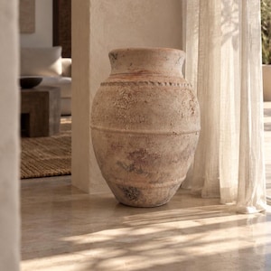 May include: A large, weathered terracotta vase with a textured, earthy surface featuring mottled shades of beige, tan, and brown. The vessel has a wide, rounded body, a distinct horizontal ridge around the neck, and a thick, flared rim opening.