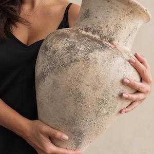 Può includere: Un grande vaso in ceramica usurata con un bordo largo e svasato e una base affusolata. La superficie presenta una finitura opaca e ruvida nei toni del beige, crema e grigio antracite, con segni di invecchiamento e una colorazione irregolare.