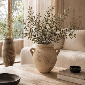 Può includere: Un vaso a urna in ceramica beige testurizzata a due manici contenente un alto ramo di ulivo frondoso con piccole olive verdi. Il vaso ha una finitura rustica e usurata e poggia su una superficie in legno, con un secondo vaso abbinato.