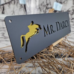 May include: A rectangular dark grey sign featuring a gold-coloured cutout of a horse jumping and the text MR. DARCY in a serif font. The sign has four circular mounting holes, one in each corner, and is displayed at an angle on a small stand.