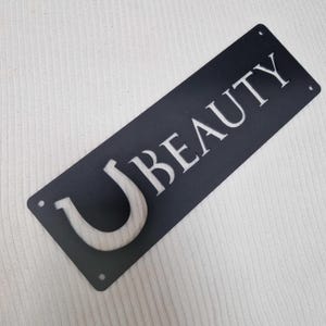 Könnte beinhalten: Ein rechteckiges schwarzes Metallschild mit dem ausgeschnittenen Wort UBEAUTY in einer fetten Serifenschrift. Das Schild verfügt über vier kleine kreisförmige Befestigungslöcher, die sich an jeder der vier Ecken befinden, zur Montage.