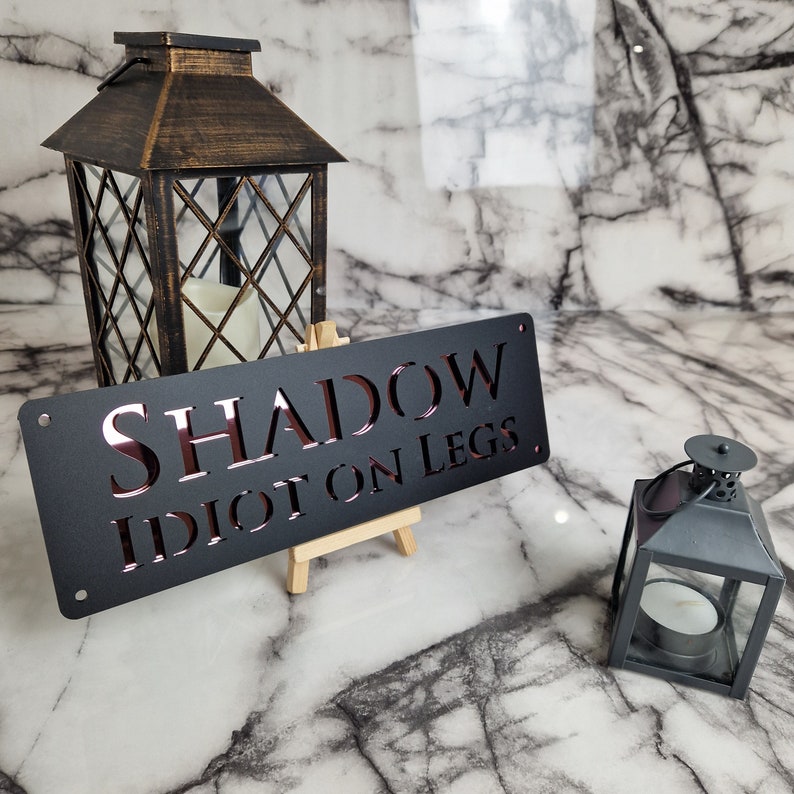 May include: Black metal sign with the text "SHADOW IDIOT ON LEGS" in a pink-red color. The sign is propped up on a small wooden easel. There is a black metal lantern with a glass window on the right side of the image.