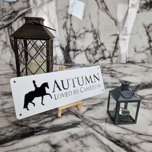 May include: White wooden sign with black text that reads "AUTUMN LOVED BY CAMERON". A black silhouette of a person riding a horse is on the left side of the sign. The sign is propped up on a small wooden easel.