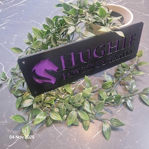 May include: A rectangular black metal sign featuring a cutout horse head silhouette and the text "HUGHIE" and "LOVED BY SASHA" with a purple backing. The sign has small circular holes at the corners for mounting and a smooth matte finish.