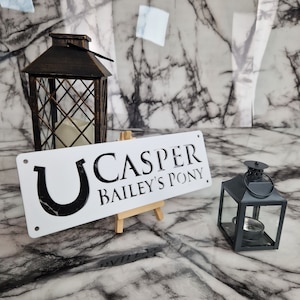 May include: White wooden sign with black text that reads "U CASPER BAILEY'S PONY". The sign is on a small wooden easel and there are two black lanterns in the background.