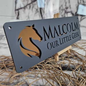 May include: A rectangular matte dark grey sign featuring a cutout of a horse head profile on the left and the text MALCOLM OUR LITTLE GEEK on the right. The cutout reveals a reflective bronze-coloured surface underneath. The sign has four holes.