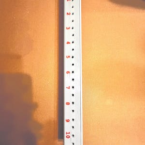 May include: A white, rectangular tool with a handle and a series of evenly spaced holes. Red numbers from 1 to 10 are printed along the side. The tool is likely used for measuring or marking.