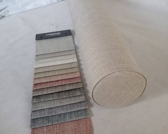 May include: A beige cylindrical pillow with a fabric cover. A fabric swatch book with a variety of colors and textures is displayed next to the pillow. The swatch book has the text "Fargotex" printed on the cover.