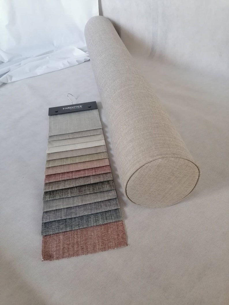 May include: A beige cylindrical pillow with a fabric cover. A fabric swatch book with a variety of colors and textures is displayed next to the pillow. The swatch book has the text "Fargotex" printed on the cover.