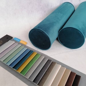 Könnte beinhalten: Zwei zylindrische Kissen in Tealblau auf weißem Hintergrund. Darunter befindet sich ein Stoffmusterbuch mit 20 verschiedenen Stoffmustern in verschiedenen Farben.