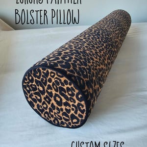 Op de afbeelding: Een cilindrisch bolsterkussen met een luipaardprint in zwart en bruin. Het kussen heeft zwarte randen aan de uiteinden. Tekst op de afbeelding luidt "Luxury Panther Bolster Pillow" en "Custom Sizes".