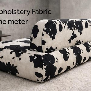 May include: A low-profile sofa upholstered in a white fabric with a black cowhide-style spotted pattern. The text Cowhide Upholstery Fabric By the meter is visible above the sofa, which features a rectangular seat and a rounded backrest.