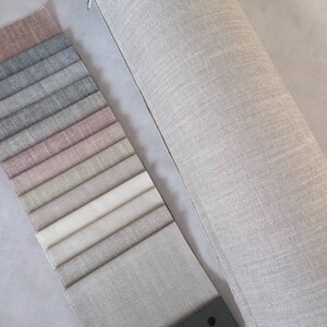 May include: A swatch book of fabric samples in various shades of gray, pink, beige, and white. The swatches are arranged in a row, with the darkest shade at the top and the lightest shade at the bottom. A roll of the same fabric in a light gray color is shown to the right of the swatches.