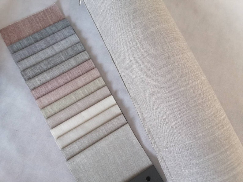 May include: A swatch book of fabric samples in various shades of gray, pink, beige, and white. The swatches are arranged in a row, with the darkest shade at the top and the lightest shade at the bottom. A roll of the same fabric in a light gray color is shown to the right of the swatches.