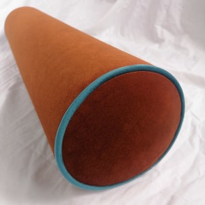 Puede incluir: Una almohada cilíndrica marrón con ribete turquesa. La almohada está hecha de una tela suave y aterciopelada.