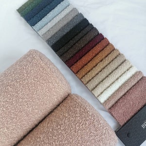 Könnte beinhalten: Eine Auswahl an Stoffmustern in verschiedenen Farben, darunter Grün, Blau, Grau und Braun, fächerförmig angeordnet. Zwei Rollen strukturierten, hellrosa Stoffes befinden sich im Vordergrund. Die Stoffe werden auf einer weißen Oberfläche präsentiert, um die Farbpalette hervorzuheben.