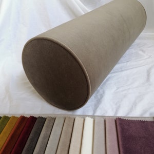May include: A brown cylindrical pillow with a soft, velvety texture. The pillow is displayed on a white surface with a variety of fabric swatches in different colors.