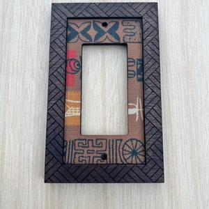 May include: A rectangular light switch cover with a dark brown, textured wooden frame. The frame surrounds a patterned fabric insert with red, blue, and tan designs. The cover has a single rectangular opening for the switch.