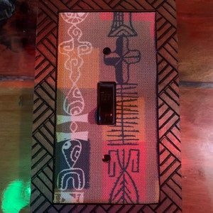 May include: A decorative light switch cover with a patterned fabric insert featuring tribal-style designs in white, blue, and yellow. The cover has a dark brown wooden frame with a geometric border and a black switch.