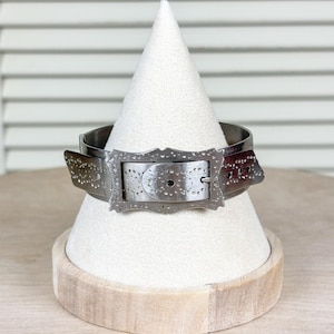 May include: A silver-coloured metal cuff bracelet with a buckle design. The bracelet features a rectangular buckle with a decorative edge and small holes. The cuff is displayed on a white, cone-shaped stand.