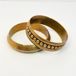 May include: Two circular brass bangles with a weathered, antique finish. One bangle features a smooth, plain surface, while the other is decorated with a raised, beaded texture running along the center of the band in a continuous pattern.