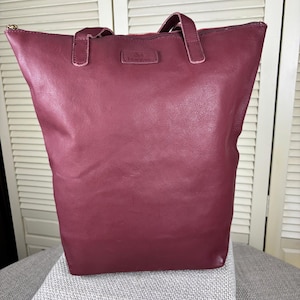 May include: A burgundy leather tote bag with a rectangular shape and two handles. The bag has a zipper closure and a small rectangular tag near the top. The bag is sitting on a gray surface.