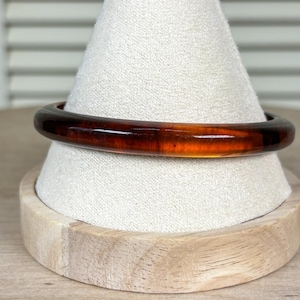 May include: A brown and amber-colored bangle bracelet. The bracelet is made of a translucent material, with a smooth, polished surface. The color transitions from dark brown to amber. The bracelet is displayed on a white fabric-covered stand.