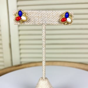May include: Gold-coloured stud earrings, each with a cluster of three coloured stones: a blue oval, a red teardrop, and a white oval. The earrings are displayed on a beige fabric earring stand.