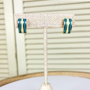 May include: A pair of gold-toned earrings with a curved, rectangular shape. Each earring features a central band of turquoise enamel, flanked by thin bands of cream enamel. The earrings are displayed on a neutral-toned jewelry stand.