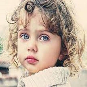 Puede incluir: Retrato de un niño pequeño con ojos azules claros y cabello rubio rizado. El niño lleva un suéter de color claro y mira directamente a la cámara. El fondo está borroso, con toques de vegetación y otros elementos indistintos.