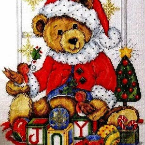 May include: A cross-stitch Christmas design showing a teddy bear in a Santa Claus outfit, with a red coat and hat trimmed with white. The bear is surrounded by presents, a Christmas tree, and blocks spelling "JOY". The colours are red, green, gold, and brown.