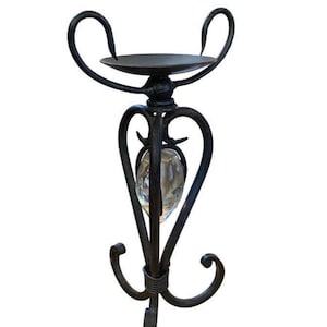 May include: A black wrought iron candle holder featuring a circular top plate, curved scrollwork arms, and a tripod base. A clear, faceted glass stone is suspended within the central metal frame, adding a reflective element to the dark metal structure.