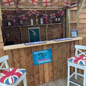Könnte beinhalten: Eine Holzbar mit einem Schild, auf dem steht "Why not? It's five o'clock somewhere!" und zwei weiße Barhocker mit rot-weiß-blauen Kissen. Die Bar ist mit Lichterketten und kleinen Flaggen geschmückt.