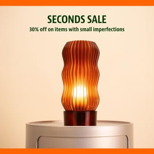 Op de afbeelding: Een gloeiende amberkleurige lamp met een geribbeld, golvend ontwerp. De lamp staat op een zilveren voet en straalt een warme gloed uit. De tekst "SECONDS SALE" en "30% korting op artikelen met kleine imperfecties" staat boven de lamp.