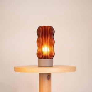 Peut inclure: Une lampe de table cylindrique dot&eacute;e d'un abat-jour ambr&eacute; translucide avec une texture verticale ondul&eacute;e et nervur&eacute;e. La lampe repose sur une base circulaire solide de couleur claire qui offre une assise stable &agrave; la structure &eacute;clair&eacute;e.