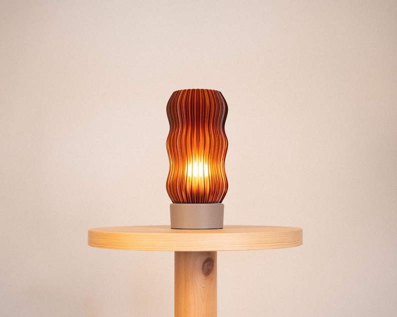 Peut inclure: Une lampe de table cylindrique dot&eacute;e d'un abat-jour ambr&eacute; translucide avec une texture verticale ondul&eacute;e et nervur&eacute;e. La lampe repose sur une base circulaire solide de couleur claire qui offre une assise stable &agrave; la structure &eacute;clair&eacute;e.