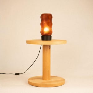 Peut inclure: Une lampe de table cylindrique avec un abat-jour brun fonc&eacute; &agrave; nervures verticales est pos&eacute;e sur une table d'appoint en bois clair. La lampe &eacute;met une lumi&egrave;re chaude. Un fin cordon d'alimentation noir part de la base de la lampe.
