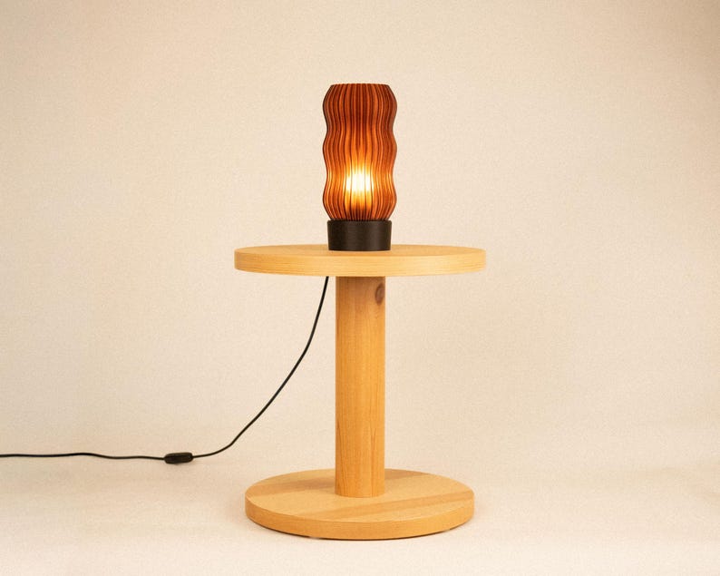 Peut inclure: Une lampe de table cylindrique avec un abat-jour brun fonc&eacute; &agrave; nervures verticales est pos&eacute;e sur une table d'appoint en bois clair. La lampe &eacute;met une lumi&egrave;re chaude. Un fin cordon d'alimentation noir part de la base de la lampe.