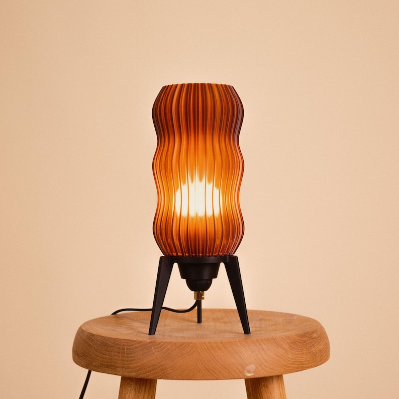 Wavy x Amber L table lamp illuminated in studio photo. Shows warm amber glow defining the wavy, textured recycled plastic shade. Retro tripod base enhances the Mid Century Modern & space age design. Eco-friendly mood light.
